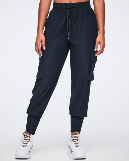 Black cargo jogger pants with oversized flap side pockets, a high-rise waistband with drawstring, and ribbed ankle cuffs, shown from the front on a female model wearing white high-top sneakers with black and leopard-print details.
