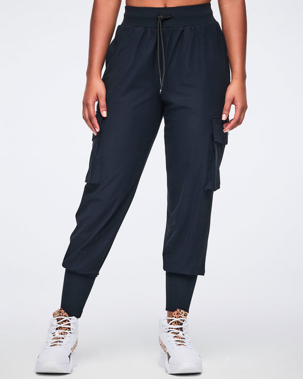 Black cargo jogger pants with oversized flap side pockets, a high-rise waistband with drawstring, and ribbed ankle cuffs, shown from the front on a female model wearing white high-top sneakers with black and leopard-print details.