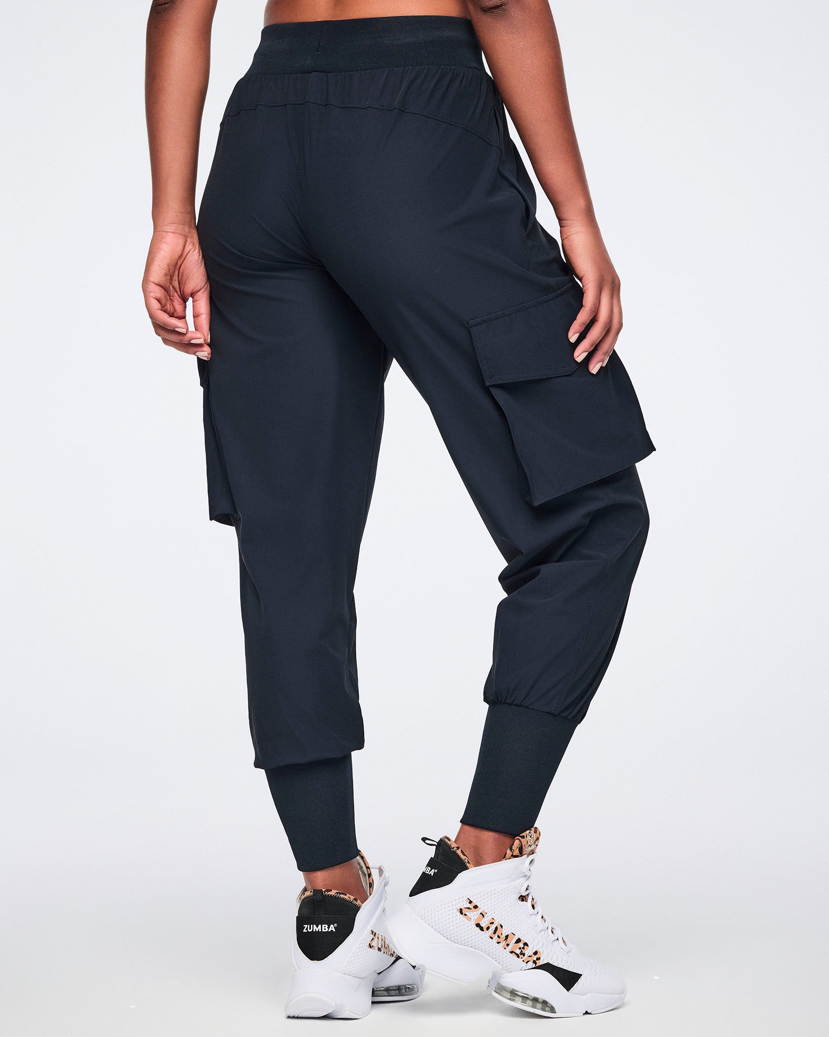 Black cargo jogger pants with oversized flap side pockets, a high-rise waistband with drawstring, and ribbed ankle cuffs, shown from the back on a female model wearing white high-top sneakers with black and leopard-print details.