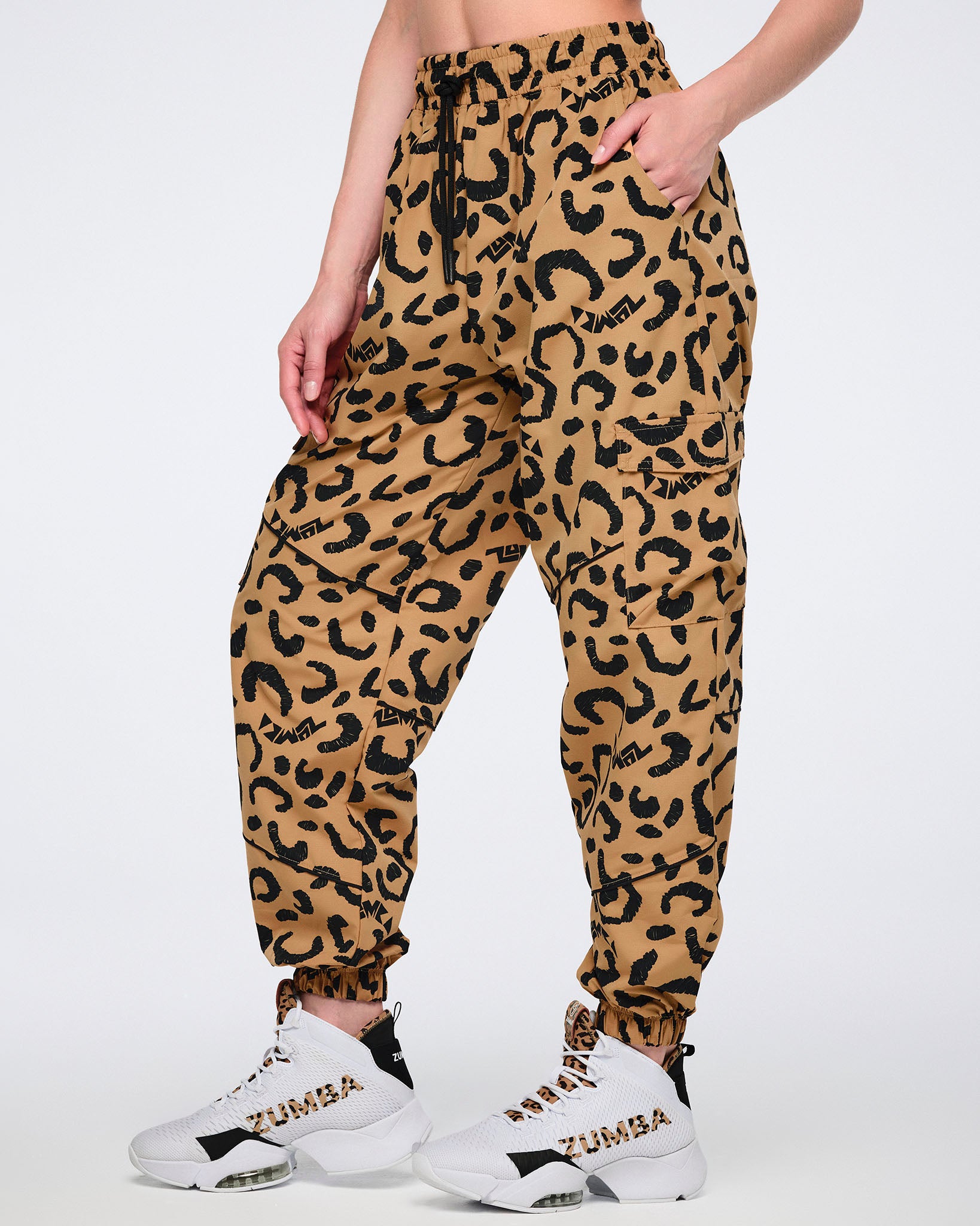 Tan leopard-print cargo pants with black abstract spots, a black drawstring waistband, flap side cargo pockets, and elastic cuffs, shown on a female model wearing white high-top Zumba sneakers with black and leopard-print accents.