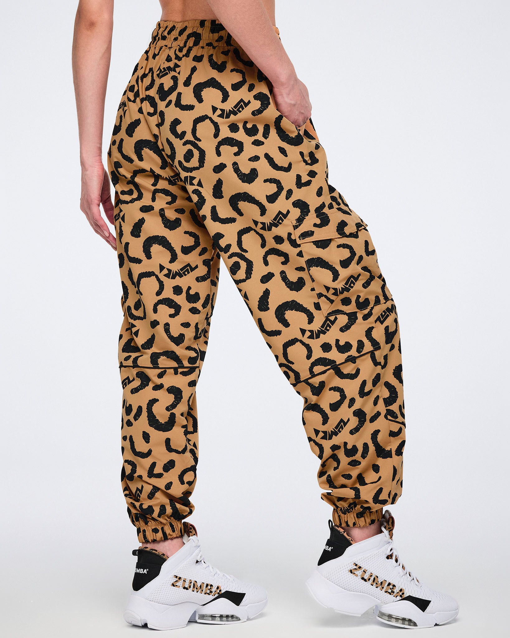 Tan leopard-print cargo pants with black abstract spots, elastic waistband and cuffs, and flap side pockets, shown from the back on a female model wearing white and black Zumba high-top sneakers with leopard-print accents.