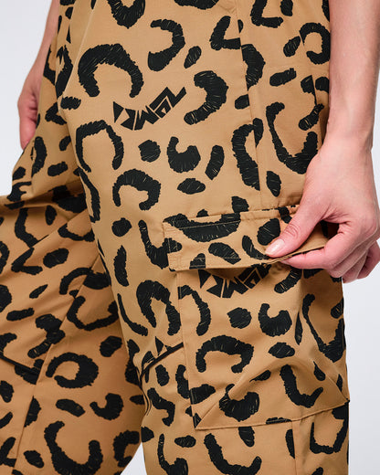 Close-up of tan leopard-print cargo pants with black abstract spots, featuring a flap side cargo pocket and logo accents, shown on a female model holding the pocket flap.