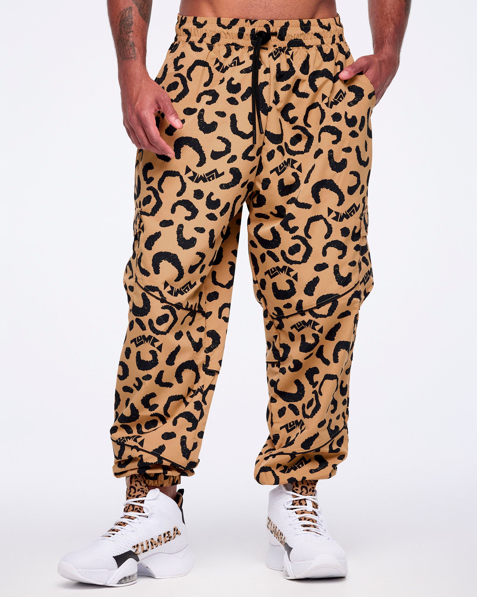 Tan leopard-print cargo pants with black abstract spots, flap side cargo pockets, a black drawstring waistband, and elastic cuffs, shown from the front on a male model wearing white high-top Zumba sneakers with leopard-print and black accents.