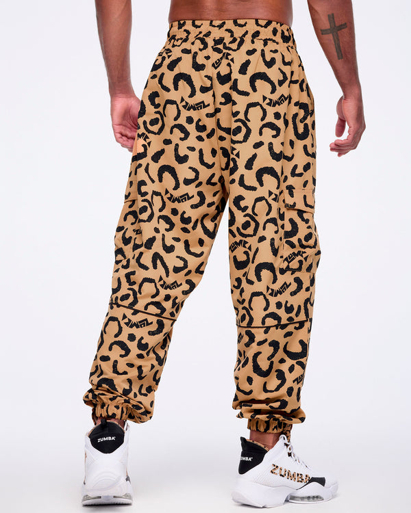 Tan leopard-print cargo pants with black abstract spots and logo accents, shown from the back on a male model wearing white high-top Zumba sneakers with black heel panels and leopard-print details.