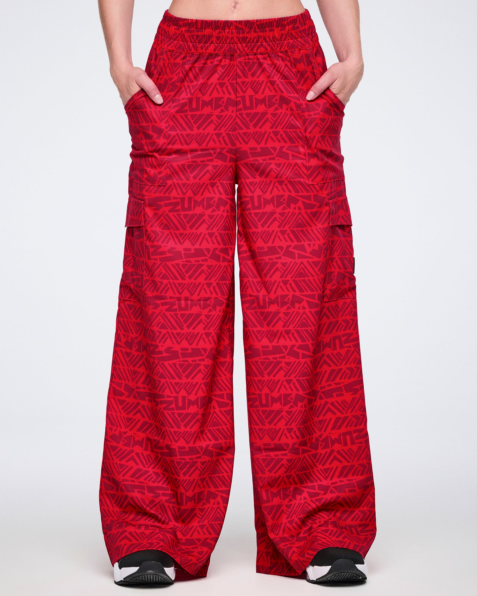 Female model wearing wide-leg red cargo pants with a tonal geometric Zumba print, front view showing a loose fit, elastic waistband, side pockets, and black athletic sneakers with white soles.