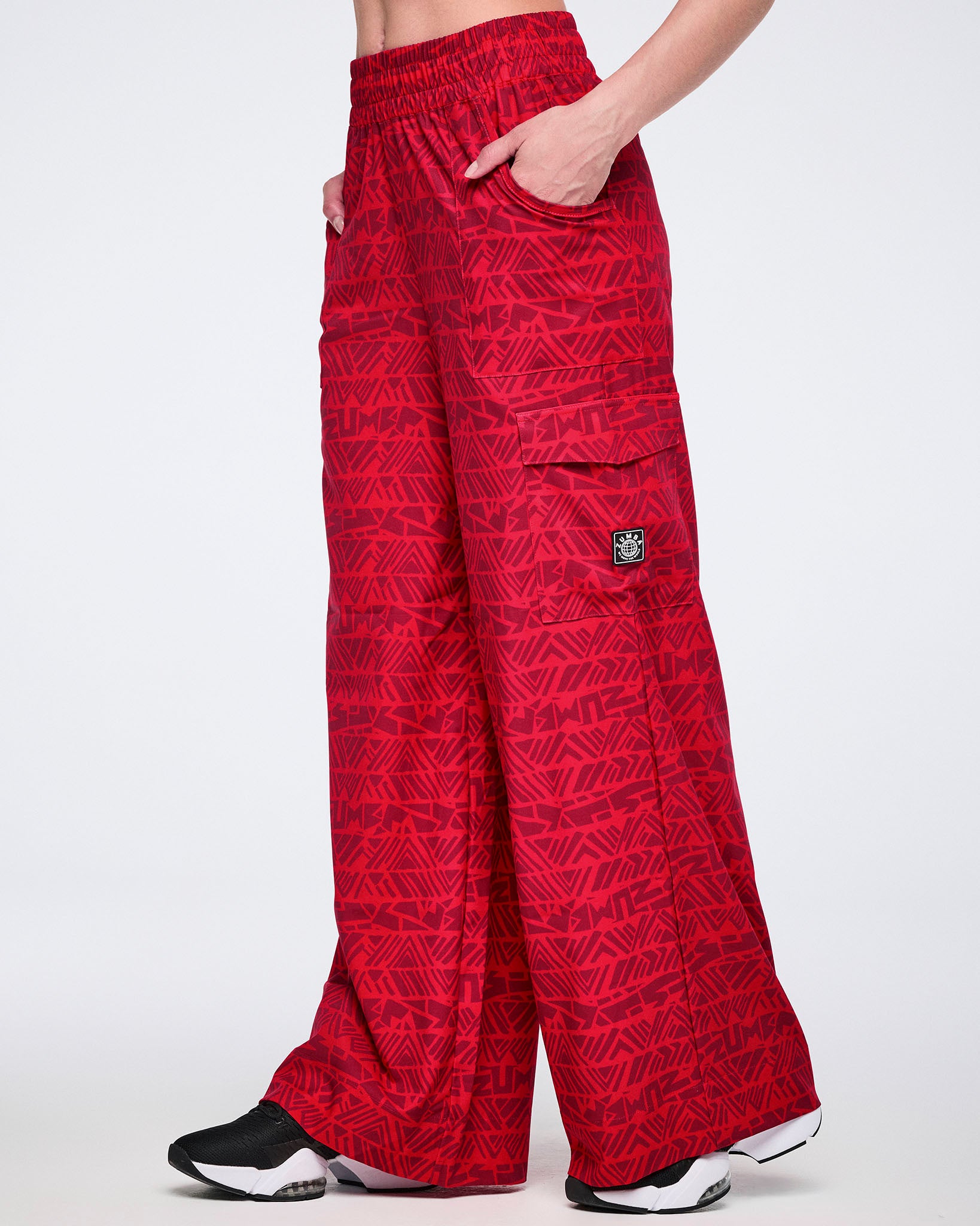 Female model wearing wide-leg red cargo pants with a tonal geometric Zumba print, elastic waistband, side pockets, flap cargo pockets, and black athletic sneakers with white soles.