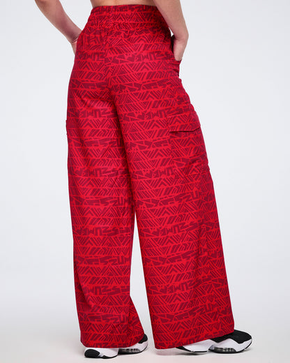 Female model wearing wide-leg red cargo pants with a tonal geometric Zumba print, shown from the back with an elastic waistband, side cargo pockets, and black athletic sneakers with white soles.