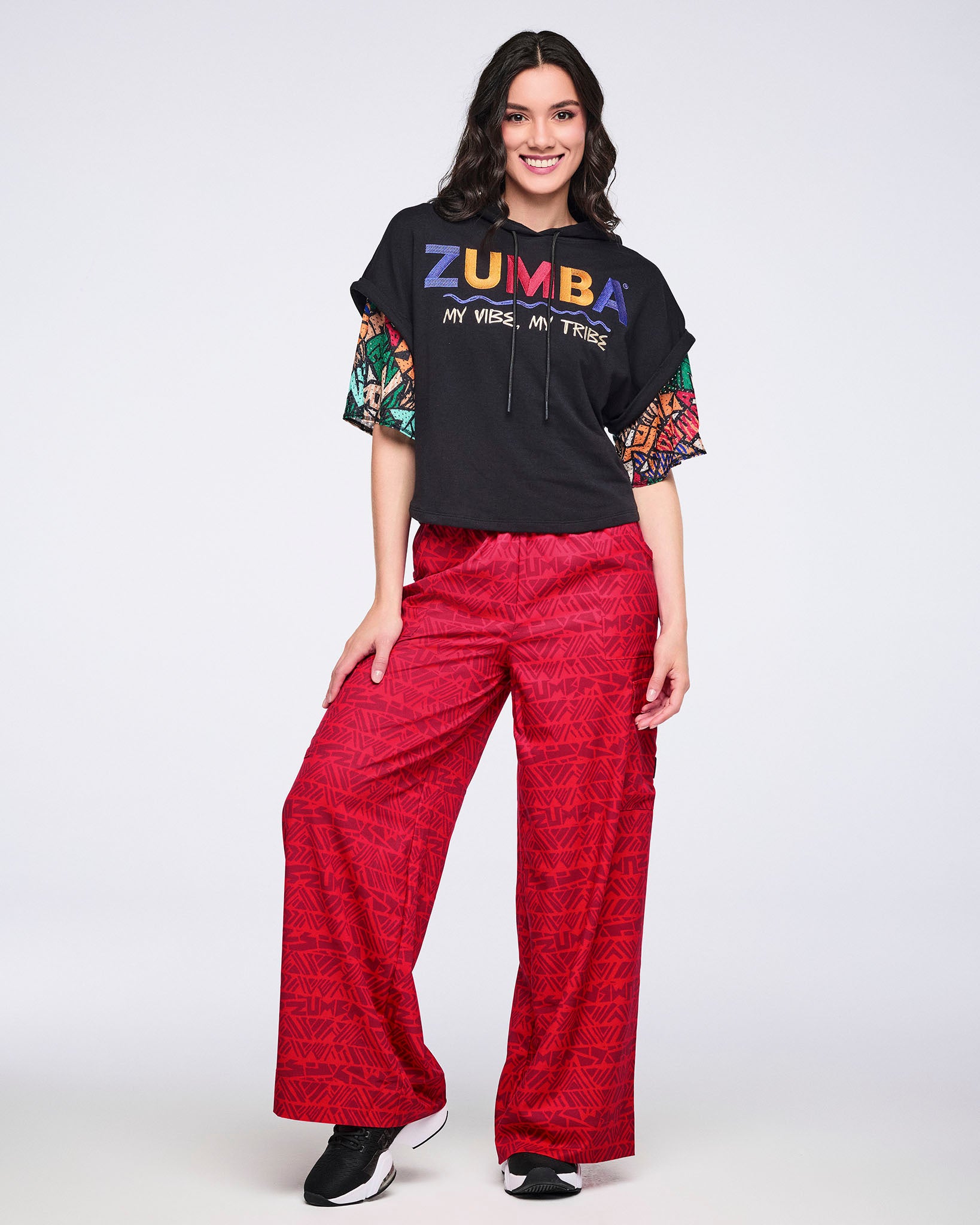 Female model wearing wide-leg red cargo pants with a tonal geometric Zumba print, paired with a black cropped hoodie with multicolor graphic lettering and a mesh patterned shirt underneath with the multicolor sleeves showing.