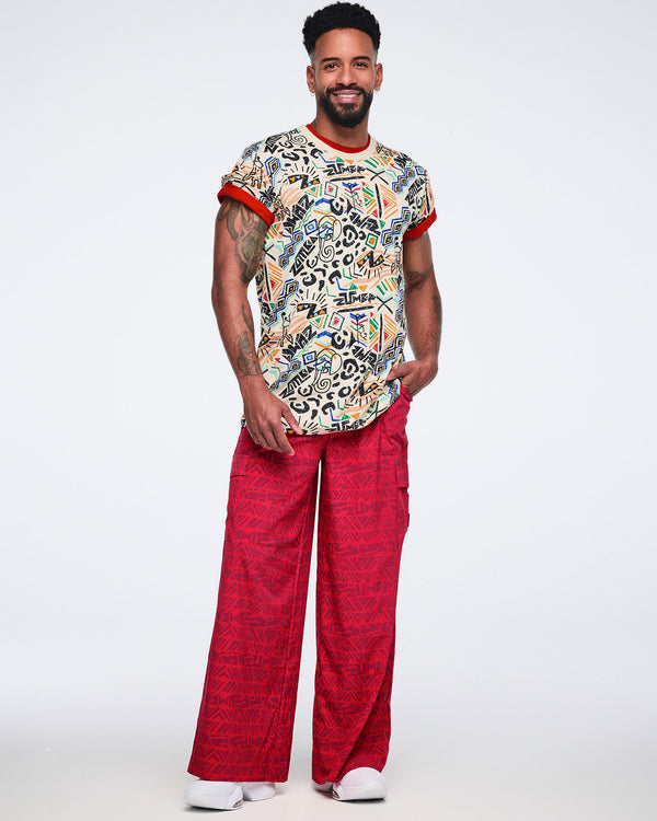 Cream short-sleeve graphic T-shirt with a crew neckline, relaxed fit, and allover multicolor abstract print featuring geometric shapes, animal-inspired motifs, and Zumba logos. Male model wears the T-shirt with red wide-leg pants.