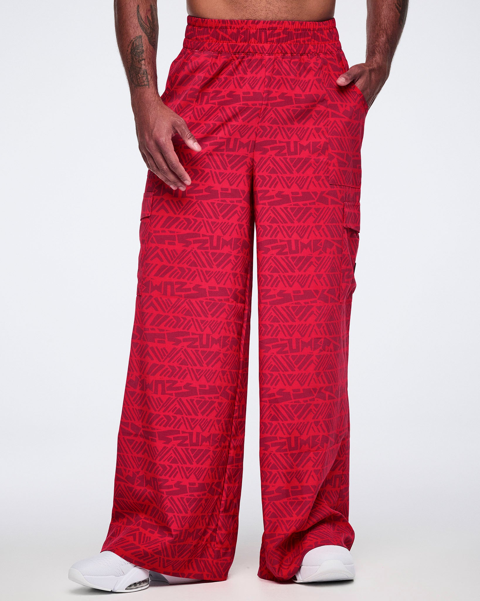 Male model wearing wide-leg red cargo pants with a tonal geometric Zumba print, front view showing a relaxed fit, side pockets, and white athletic sneakers.
