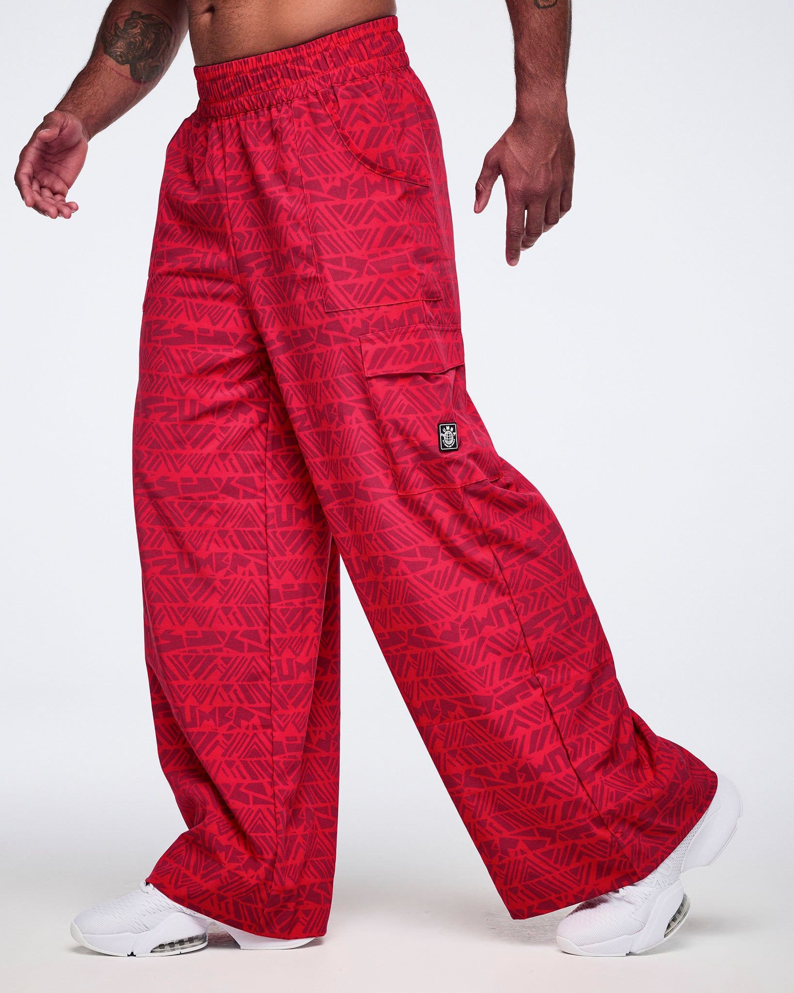 Male model wearing wide-leg red cargo pants with a tonal geometric Zumba print, side view showing the loose fit, flap cargo pocket, and white athletic sneakers.