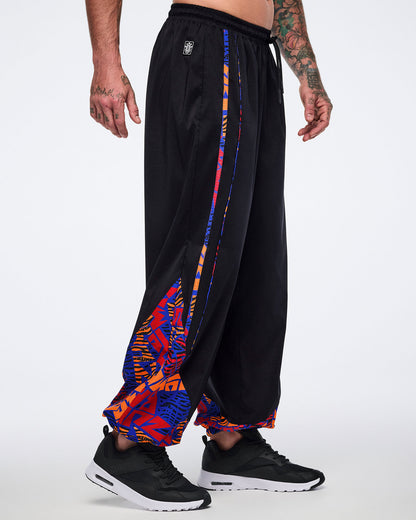 Side view of black loose-fit dance pants with an elastic drawstring waistband, side pockets, and blue, orange, and red abstract print trim and lower-leg panels. Male model wears the pants with black athletic sneakers.