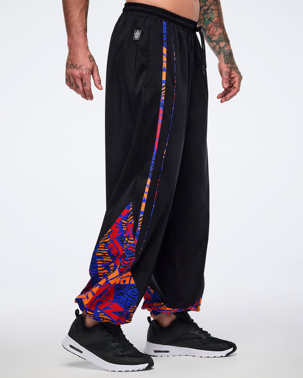 Side view of black loose-fit dance pants with an elastic drawstring waistband, side pockets, and blue, orange, and red abstract print trim and lower-leg panels. Male model wears the pants with black athletic sneakers.