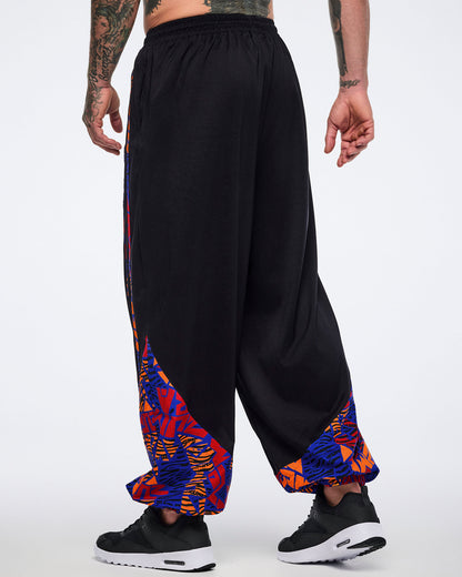 Back view of black loose-fit dance pants with a draped silhouette, elastic waistband, and blue, orange, and red abstract print panels at the lower legs. Male model wears the pants with black athletic sneakers.