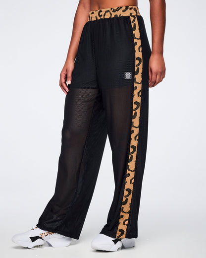 Black mesh wide-leg pants with a sheer finish, tan leopard-print waistband, matching leopard-print side stripes, and a small front logo patch, shown on a female model wearing white sneakers.