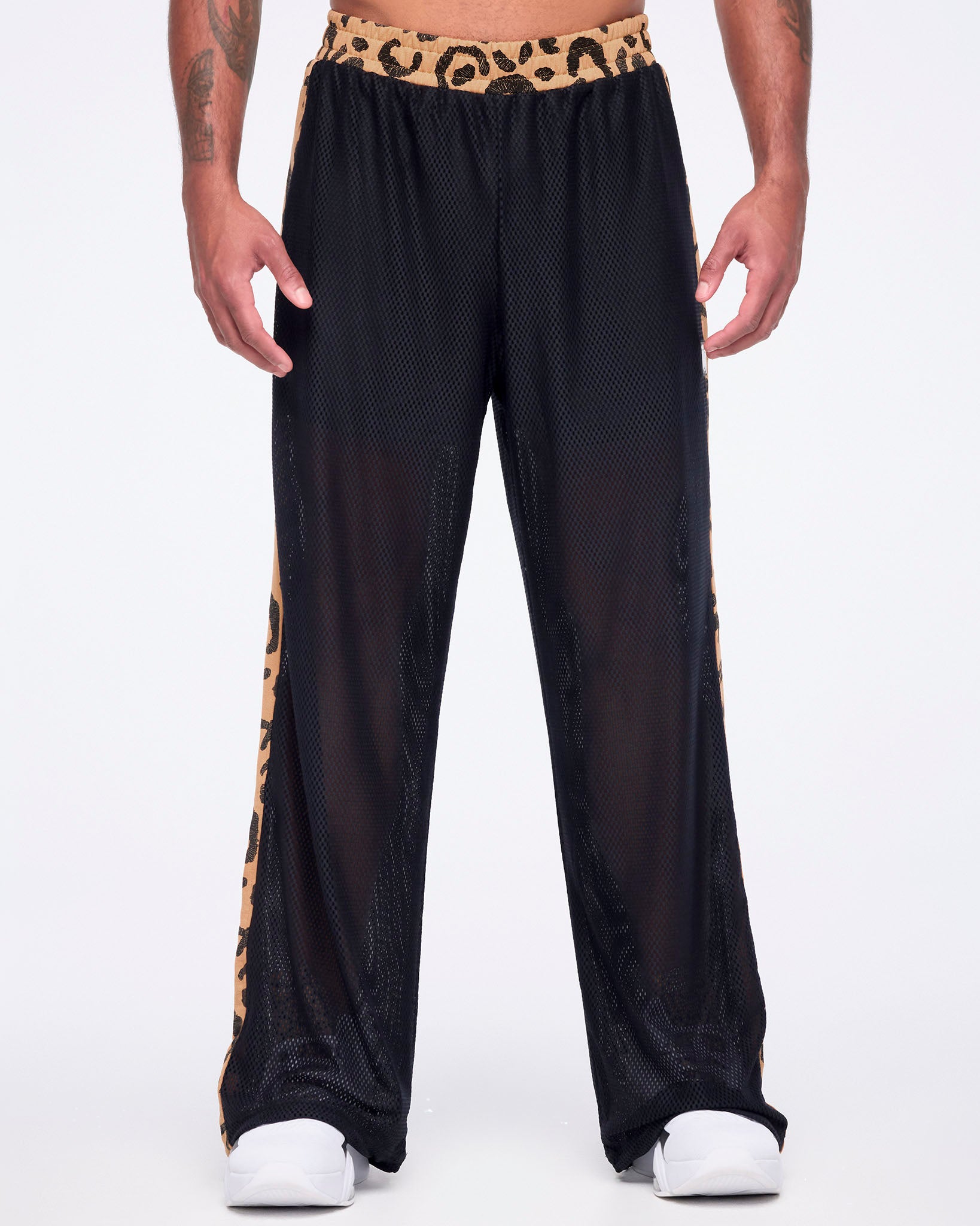 Black mesh wide-leg pants with a sheer finish, tan leopard-print waistband, and matching leopard-print side stripes, shown from the front on a male model wearing white sneakers.