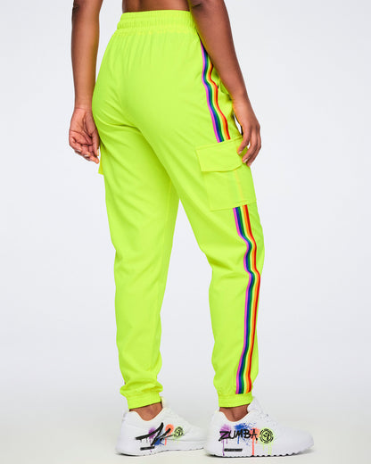 A female model wearing bright yellow pants with a rainbow stripe running down the leg and a side cargo pocket, paired with white graffiti-style sneakers, shown from the back against a plain white background.