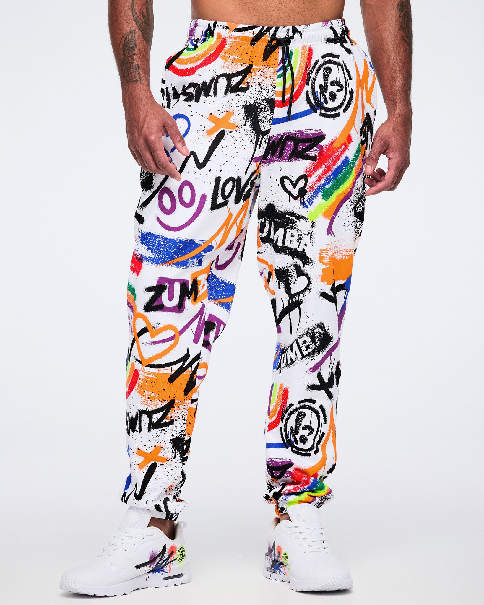 Male model wearing colorful graffiti-style pants with white Zumba shoes on a white background.
