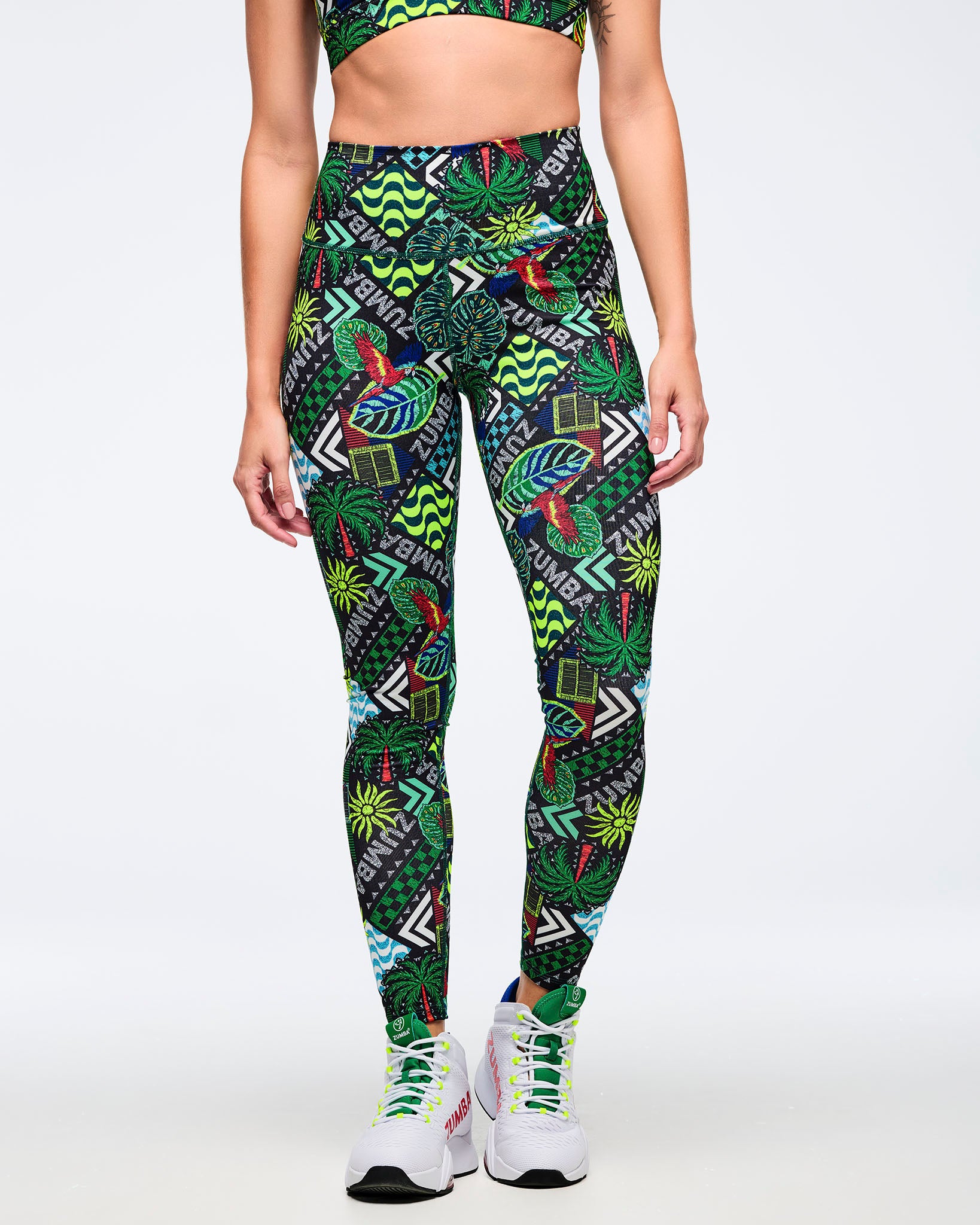 High-waisted Zumba leggings with bold tropical patchwork print featuring parrots, palm leaves, wave graphics, and “ZUMBA” text in green, teal, black, and red. Female model wears the leggings with a matching printed sports bra and white Zumba high-top sneakers on a light gray studio background.
