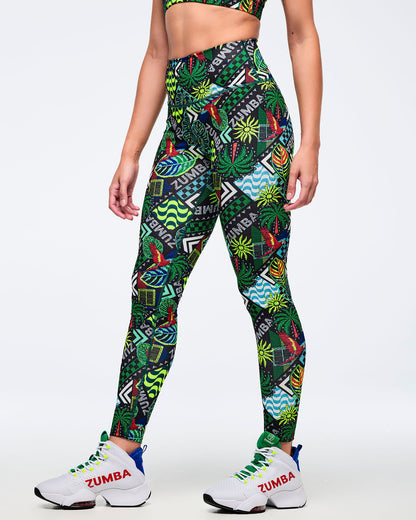 Side view of high-waisted Zumba leggings in a multicolor tropical patchwork print with “ZUMBA” lettering and geometric panels. Female model pairs them with white Zumba high-top sneakers with red logo white shoe laces and neon lace details on a light gray studio background.