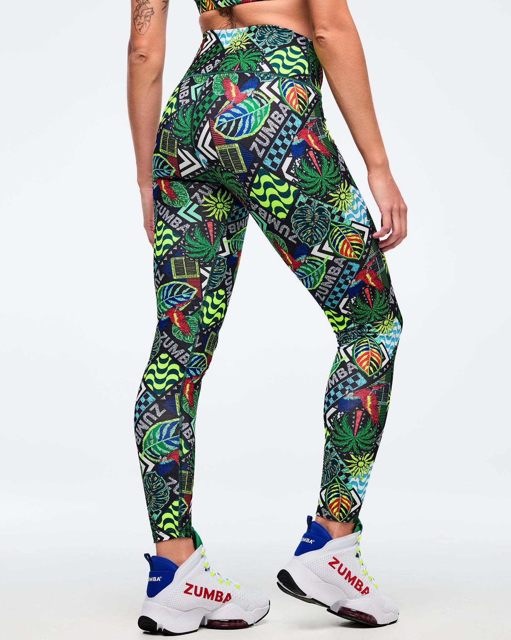 Back view of high-waisted Zumba leggings with tropical patchwork print and “ZUMBA” text accents, showing a smooth, fitted silhouette. Female model wears white Zumba high-top sneakers on a light gray studio background.
