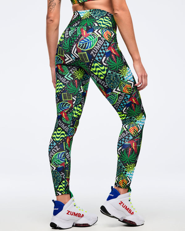 Back view of high-waisted Zumba leggings with tropical patchwork print and “ZUMBA” text accents, showing a smooth, fitted silhouette. Female model wears white Zumba high-top sneakers on a light gray studio background.