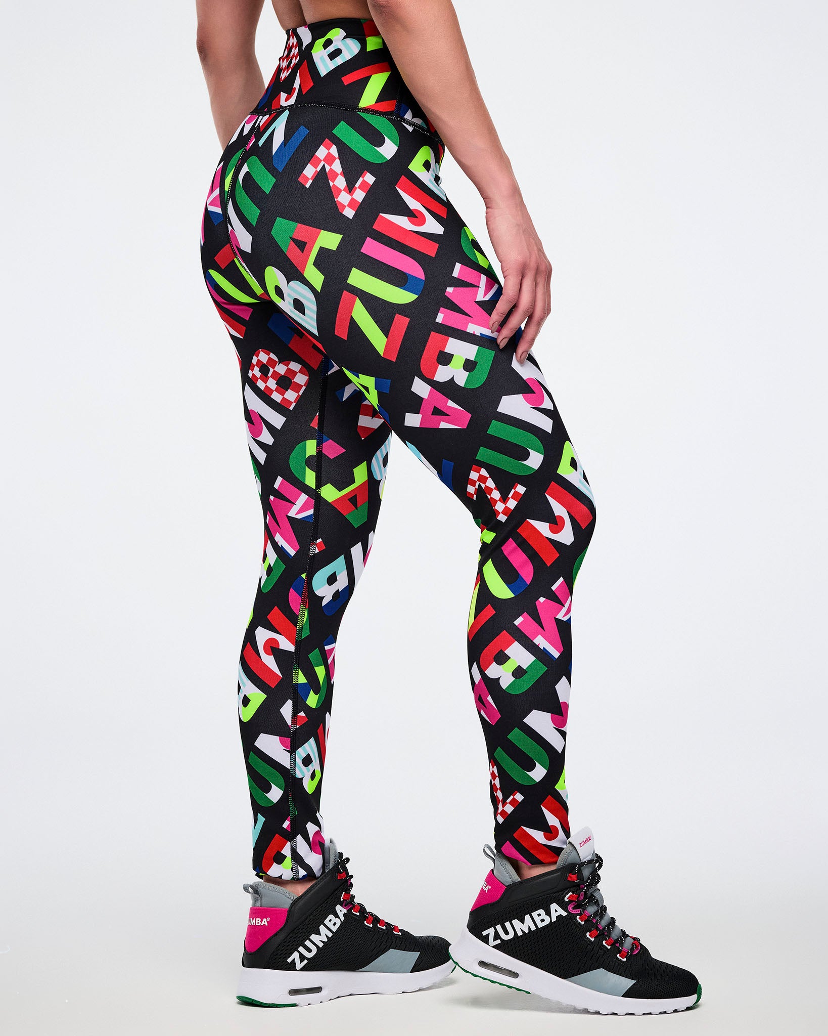 Side and partial back view of a female model wearing leggings with colorful Zumba flag-like pattern on a white background.