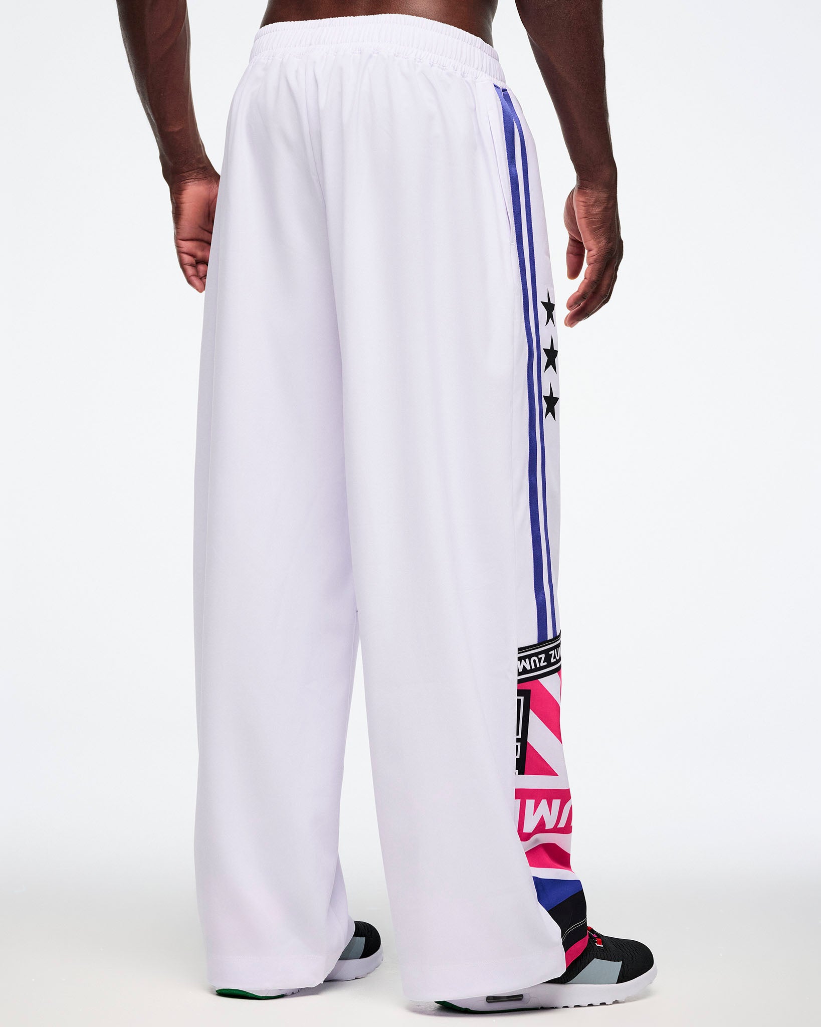 Back view of a male model in white pants with colorful side stripes and Zumba patterns, all on a white backgrounds.