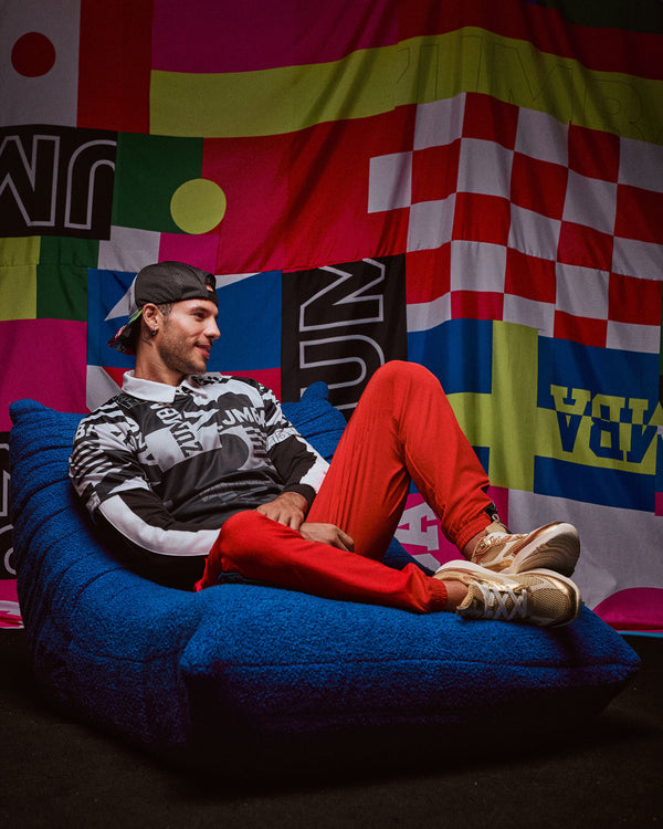 Male model wearing a backward baseball cap, seated on a blue bean bag in front of a colorful abstract wall, dressed in a black-and-white shirt, red cargo pants, and gold shoes.