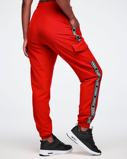Back view of a female model wearing red cargo pants with black and white stripes saying 'Zumba' in white on a white background.