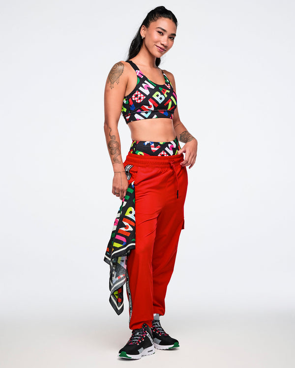 Female model wearing a colorful sports bra and red pants with a patterned bandana, on a white background.