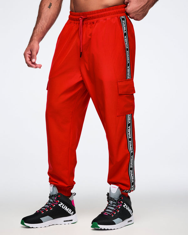 Male model wearing red cargo pants with black and white stripes saying 'Zumba' in white, paired with black and pink shoes on a white background.