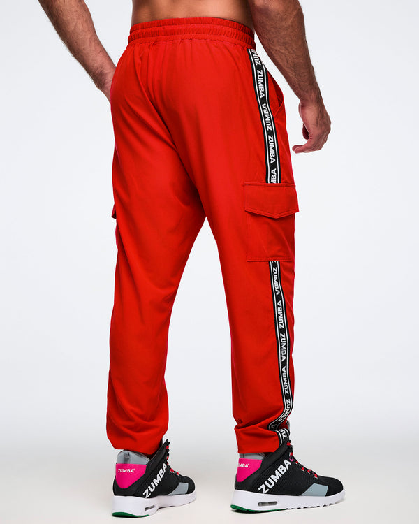 Back view of a male model wearing red cargo pants with black and white stripes saying 'Zumba' in white, paired with black and pink shoes on a white background.