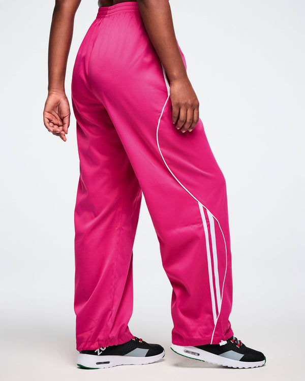 Back view of a female model wearing bright pink track pants with white stripes on a white background.
