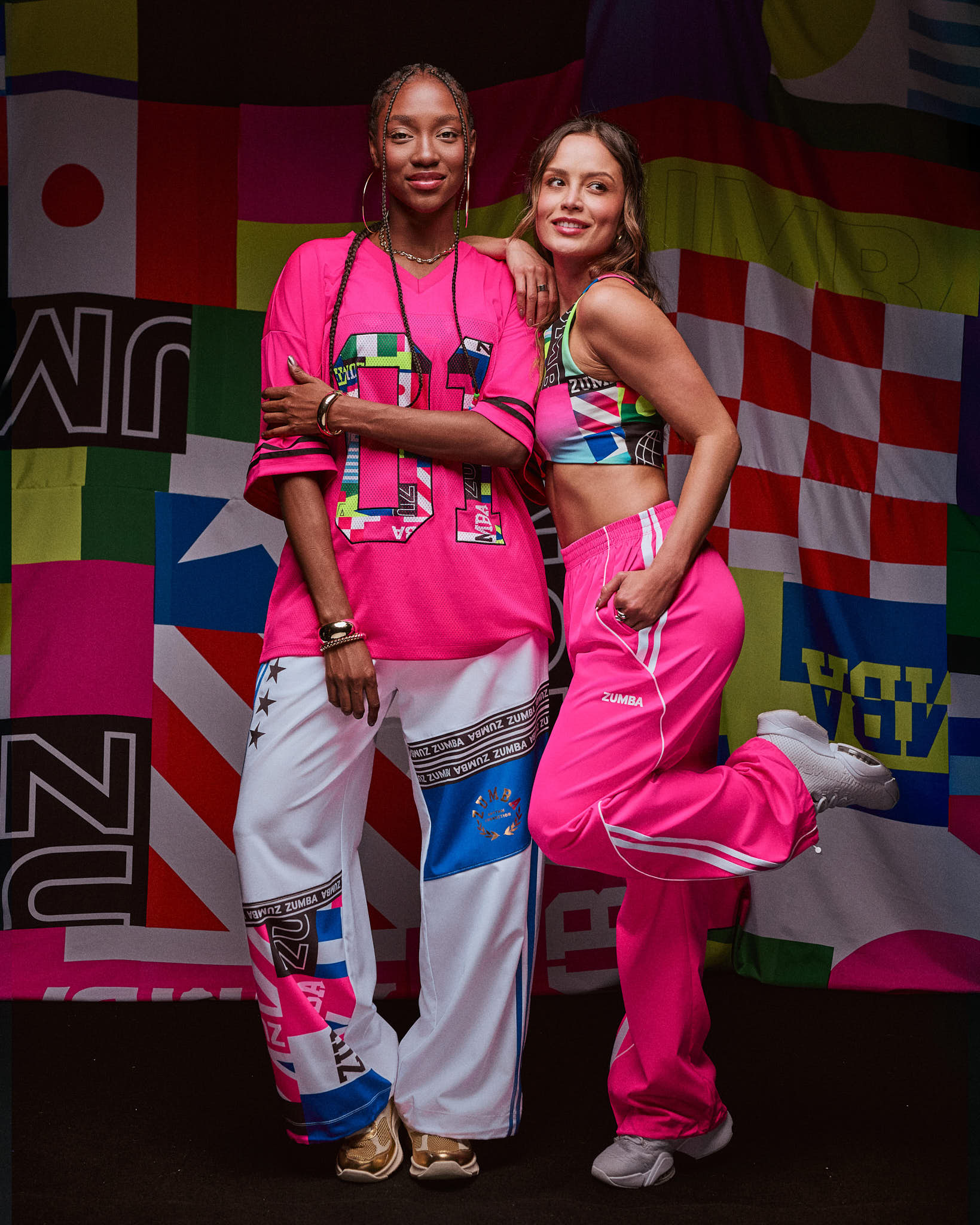 Two female models posing together in colorful outfits against a vibrant, patterned background.
