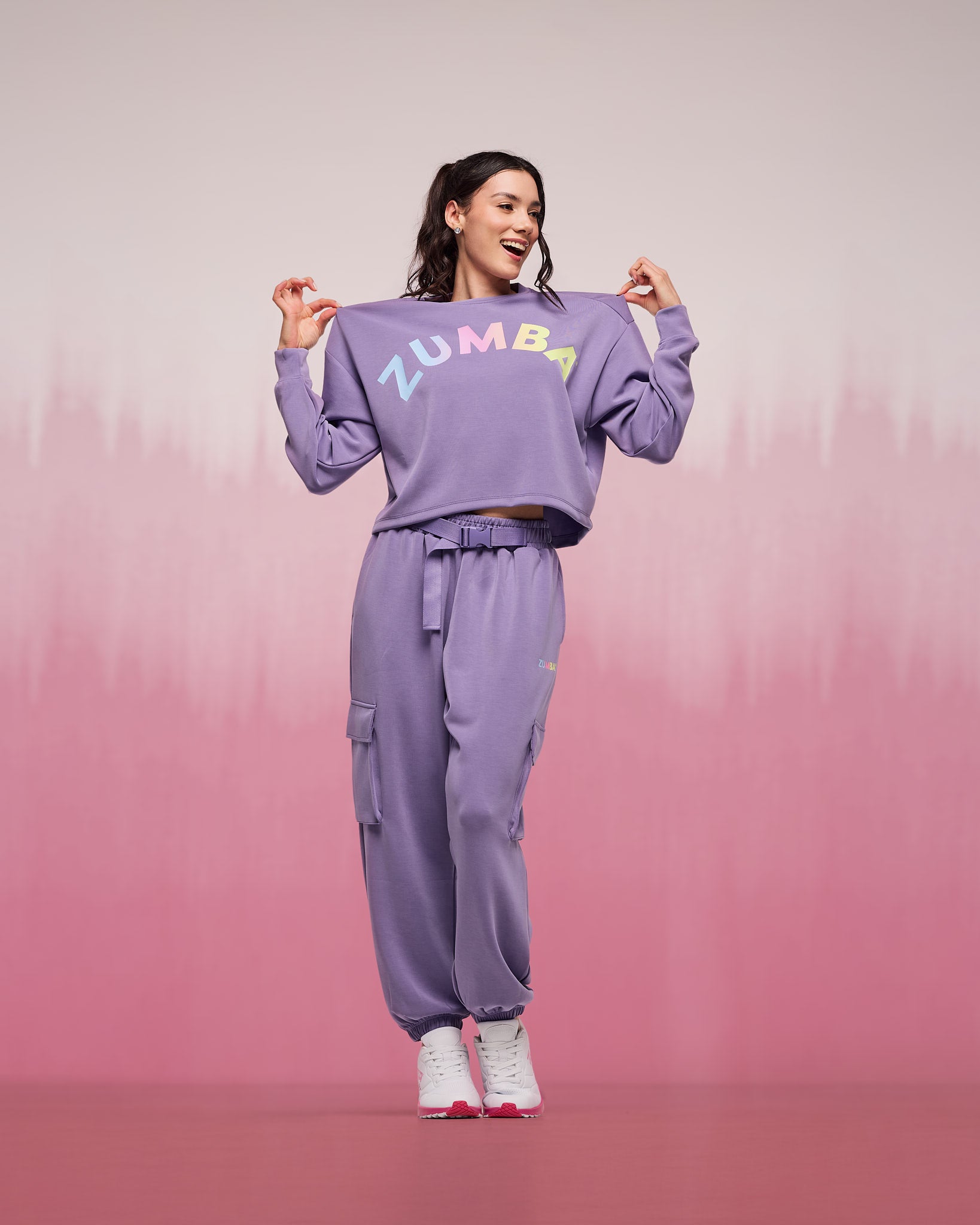 Matching lilac sweatsuit featuring a cropped crewneck sweatshirt with rainbow “ZUMBA” lettering and high-waisted cargo jogger pants with flap pockets and ankle cuffs, worn by a female model with white sneakers against a pink gradient background.
