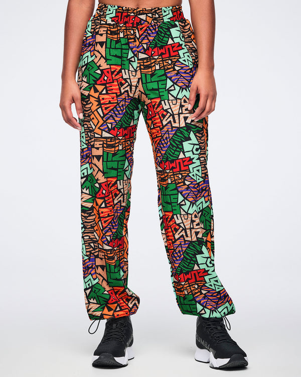 Front view of multicolor Zumba print track pants with an elastic waistband, relaxed straight-leg fit, side pockets, and adjustable ankle bungee cords. Female model wears the pants with black athletic sneakers.