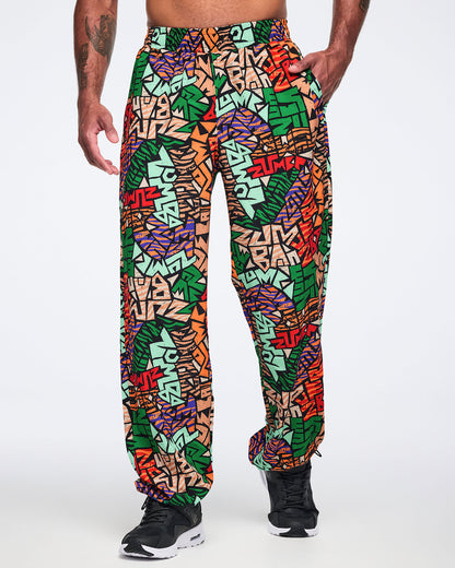 Front view of multicolor Zumba print track pants with an elastic waistband, side pockets, relaxed fit, and gathered ankle cuffs. Male model wears the pants with black athletic sneakers.