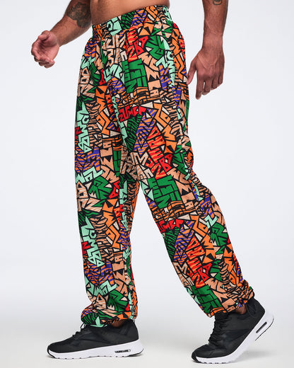 Side view of multicolor Zumba print track pants with an elastic waistband, relaxed fit, and gathered ankle cuffs. Male model wears the pants with black athletic sneakers.