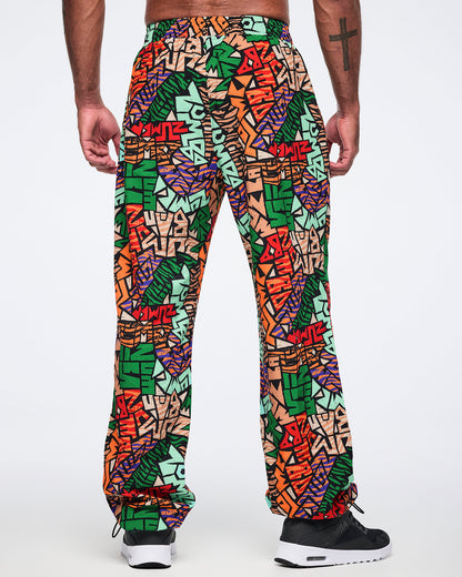 Back view of multicolor Zumba print track pants with an elastic waistband, relaxed fit, and gathered ankle cuffs. Male model wears the pants with black athletic sneakers.