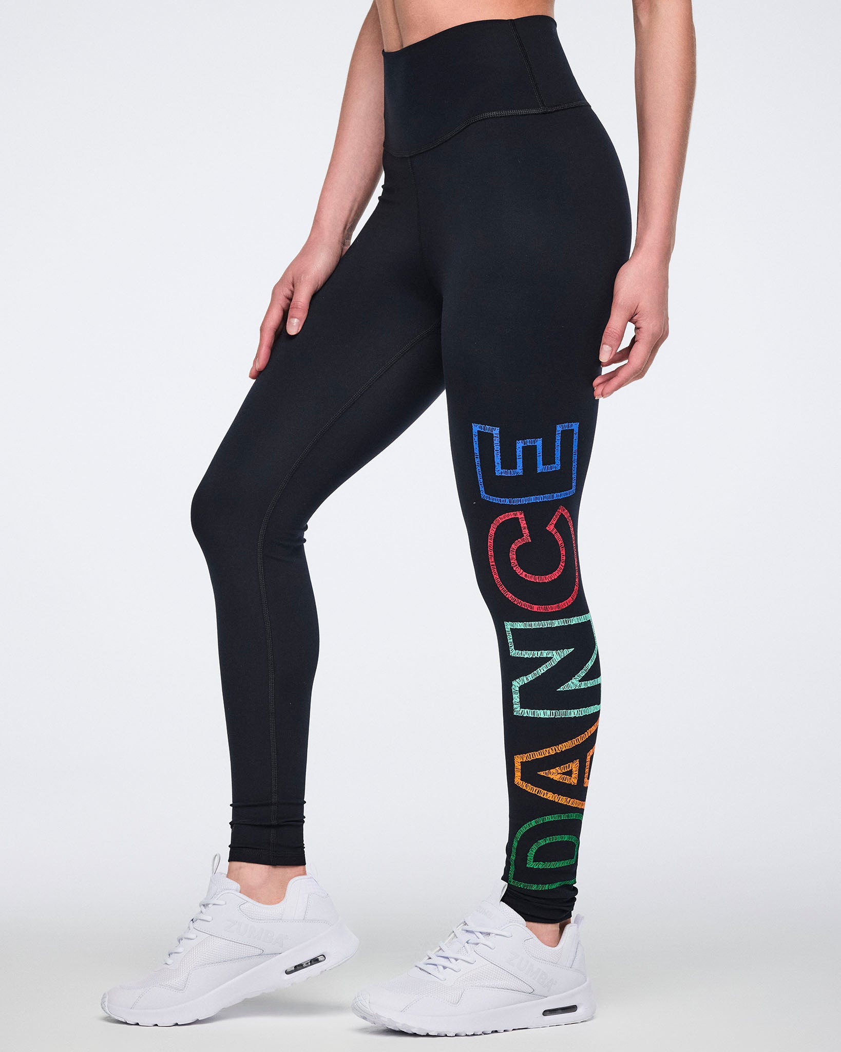Side view of black high-waisted full-length leggings with a fitted silhouette and multicolor vertical “DANCE” graphic on one leg. Female model wears the leggings with white athletic sneakers.