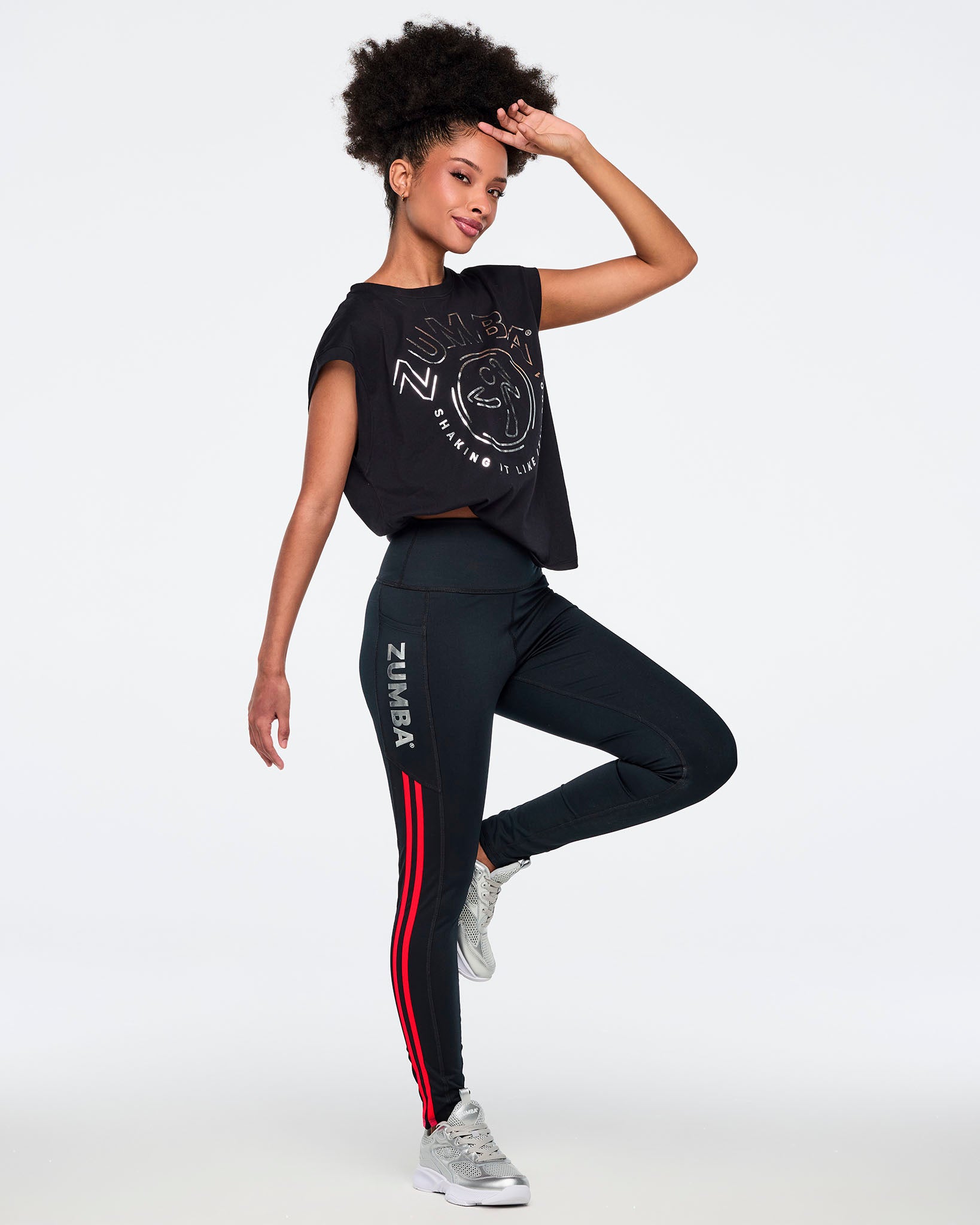 Black high-waisted leggings with silver vertical Zumba logo and bright red double side stripes. Female model wears fitted full-length leggings with a wide waistband, a black cropped Zumba graphic top, and silver athletic sneakers, posing in a full-body side view against a light gray background.