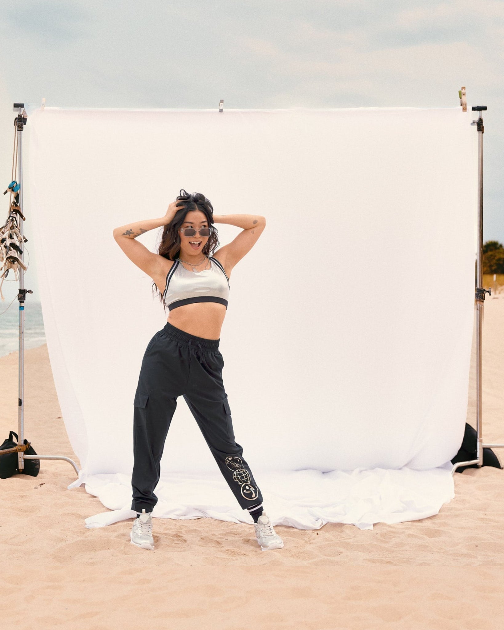 Black cargo jogger pants with a silver Zumba graphic on the lower leg. Female model wears the black joggers with a metallic silver sports bra, white sneakers, standing on sand in front of a white backdrop at the beach.