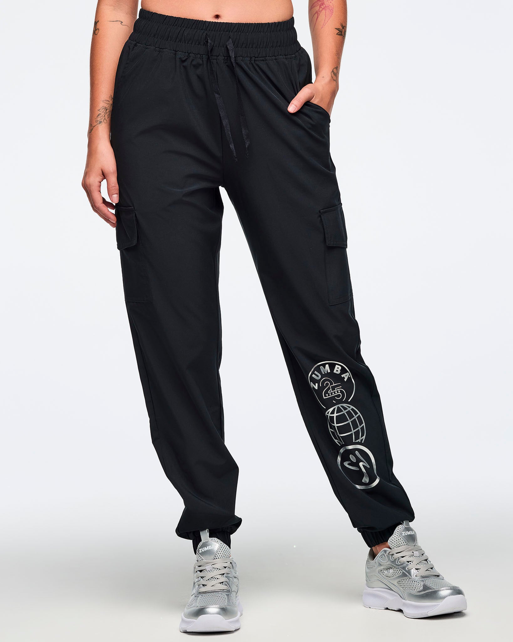 Black jogger pants with an elastic drawstring waistband, cargo pockets, and a silver Zumba 25 Years globe graphic on the lower leg. Female model is wearing the tapered joggers with silver athletic sneakers.