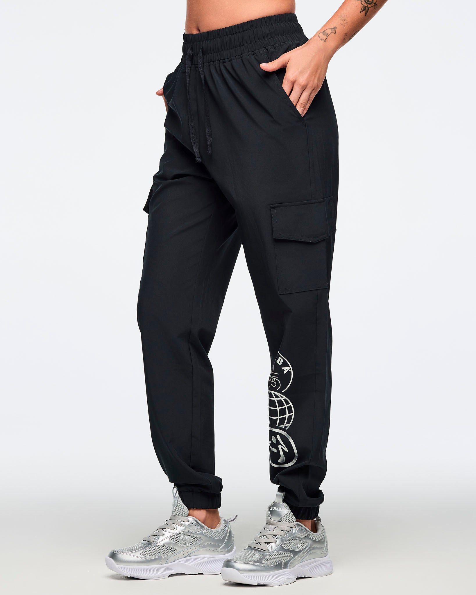 Black jogger pants with an elastic drawstring waistband, cargo pockets, and a silver Zumba 25 Years globe graphic on the lower leg. Female model is wearing the tapered joggers with silver athletic sneakers.