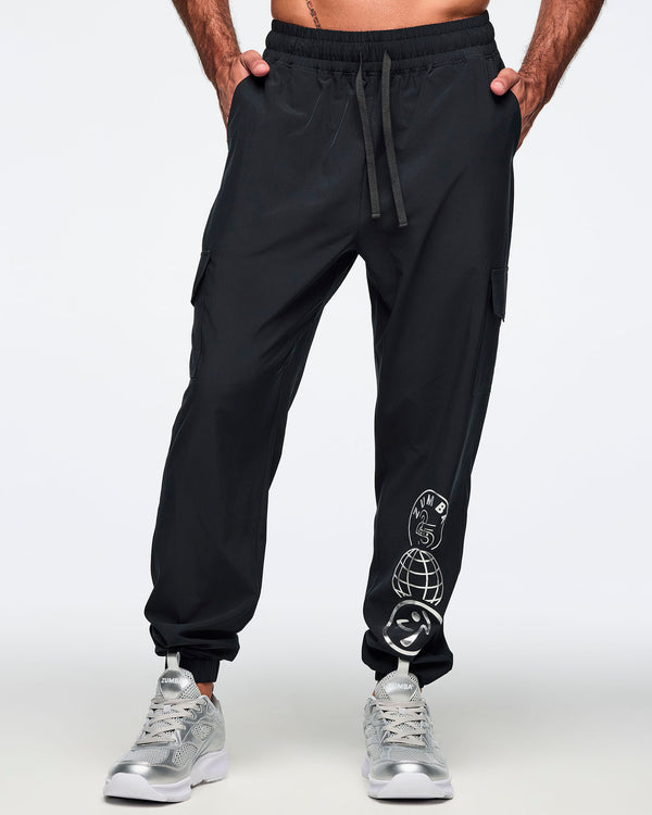 Black jogger pants with an elastic drawstring waistband, cargo pockets, and a silver Zumba 25 Years globe graphic on the lower leg. Male model is wearing the tapered joggers with silver athletic sneakers.