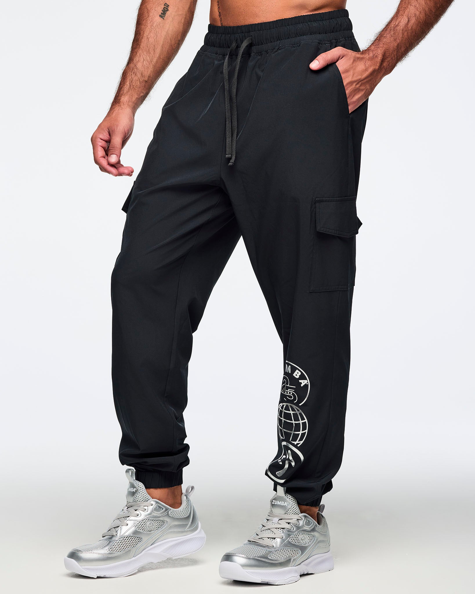 Black jogger pants with an elastic drawstring waistband, side pockets, cargo pockets, and a silver Zumba 25 Years globe graphic on the lower leg. Male model is wearing the tapered joggers with silver athletic sneakers.