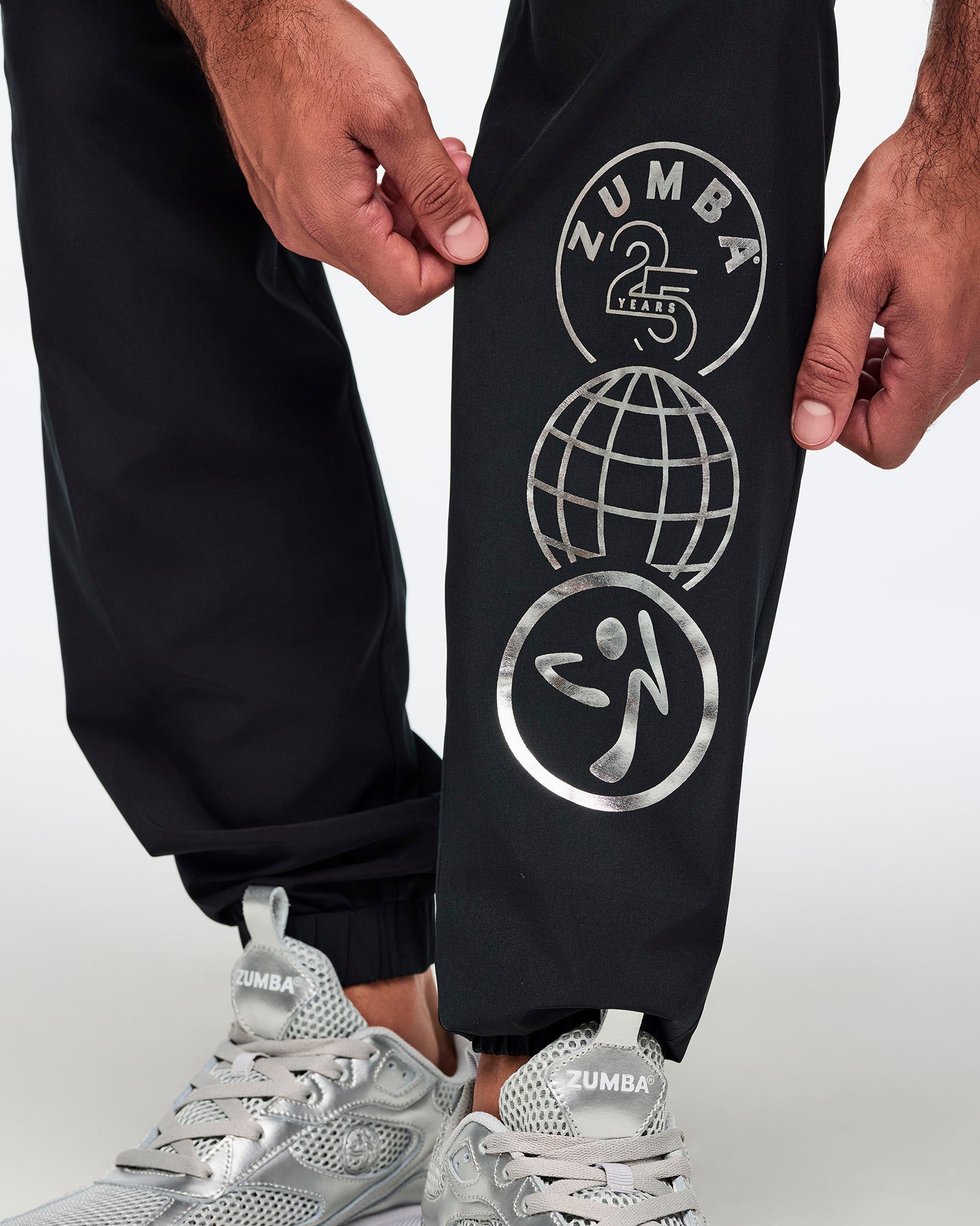 Black jogger pants with a silver Zumba 25 Years globe graphic on the lower leg. Male model is shown in a close-up view highlighting the printed leg detail and silver athletic sneakers.