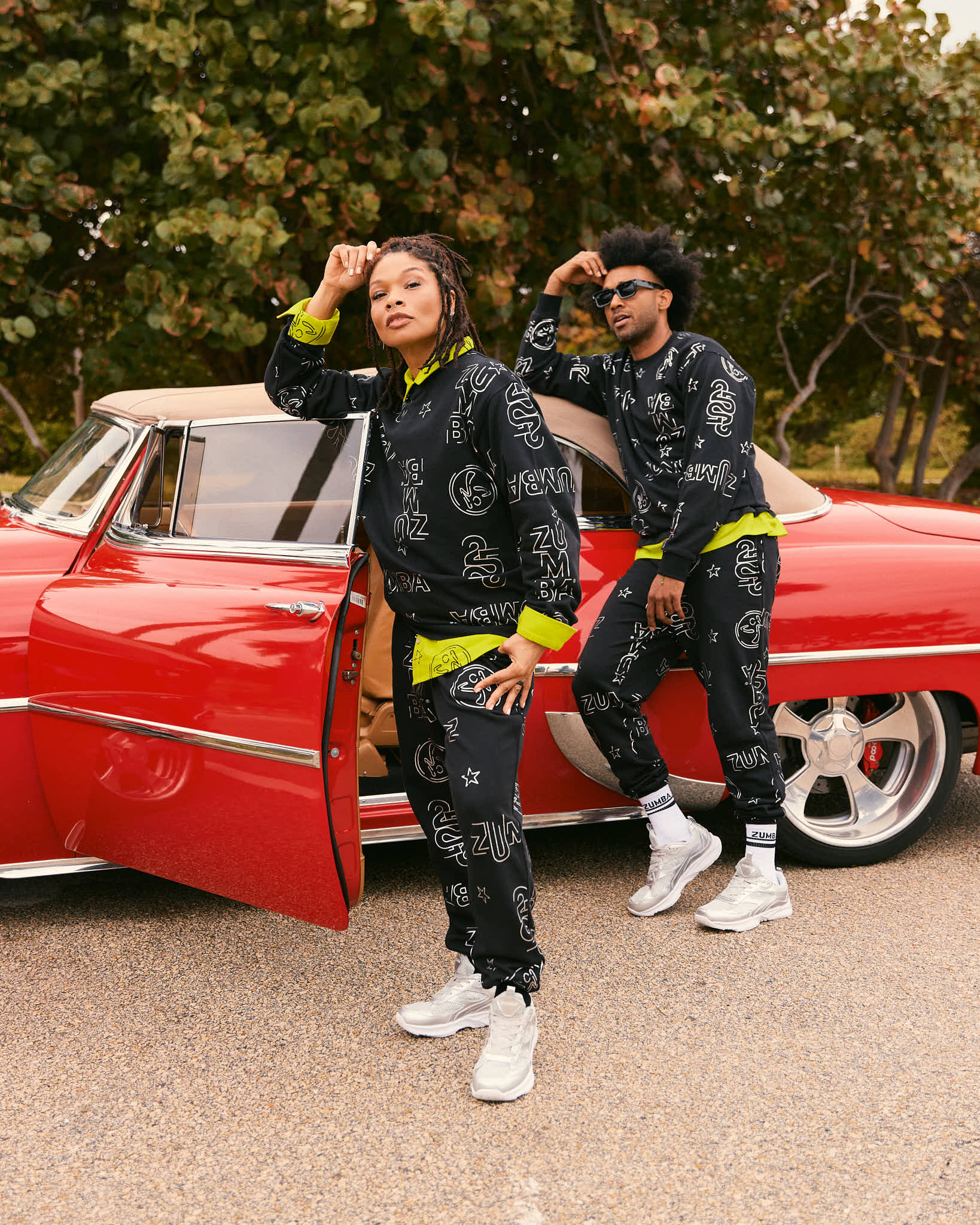 Black Zumba matching set with allover silver logo print and neon yellow trim. Female model wears a black long-sleeve sweatshirt and jogger pants with silver sneakers, while a male model wears the same printed sweatshirt and joggers with white socks and silver sneakers beside a red vintage car.