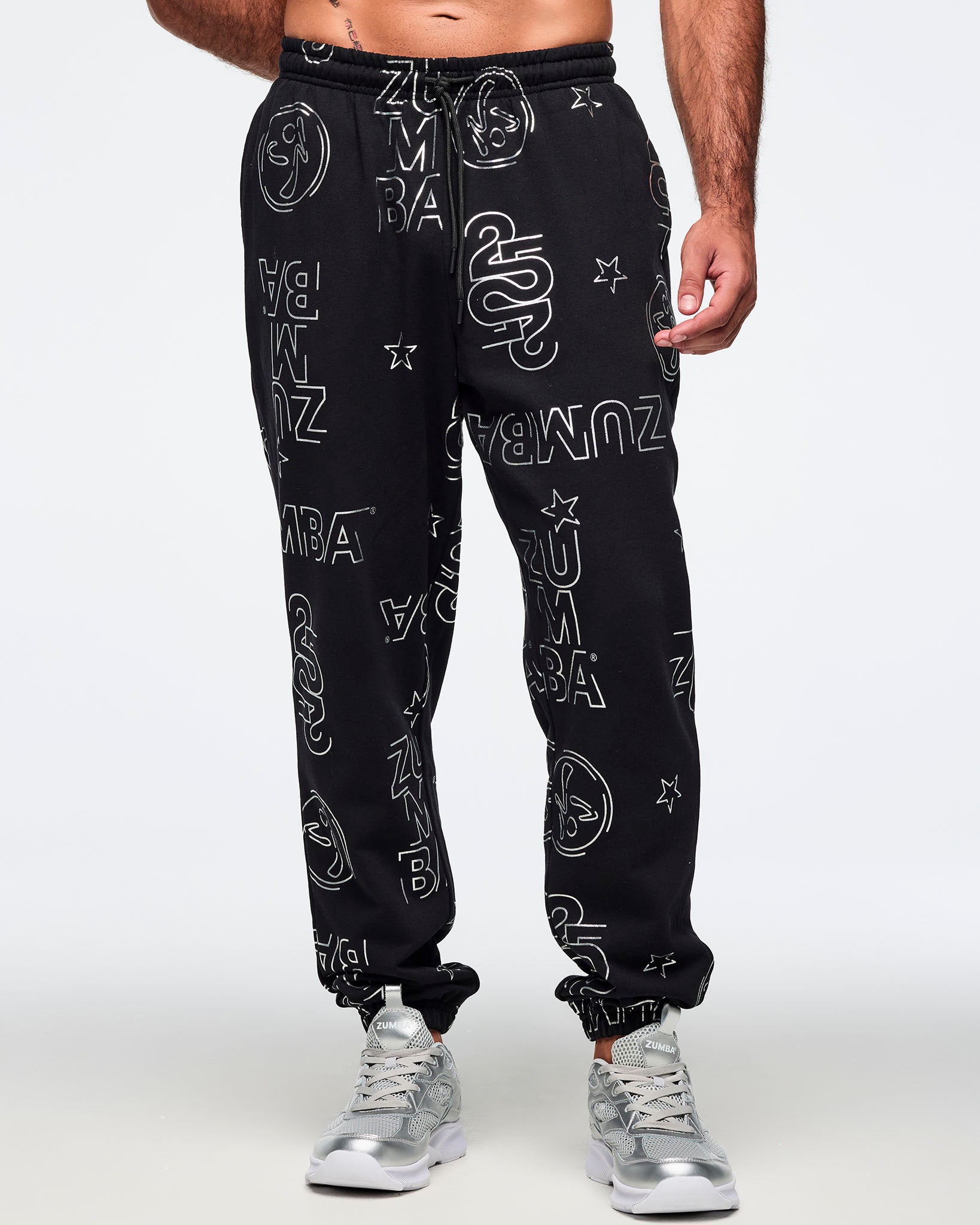 Black jogger sweatpants with an allover silver Zumba logo, icon, and star print, plus an elastic drawstring waistband and cuffed ankles. Male model is wearing the relaxed-fit joggers with silver athletic sneakers.